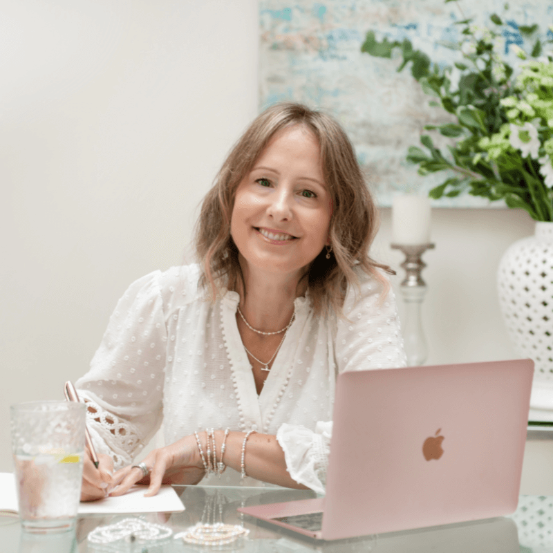 Woman sitting at a table with a pink laptop, smiling at the camera.