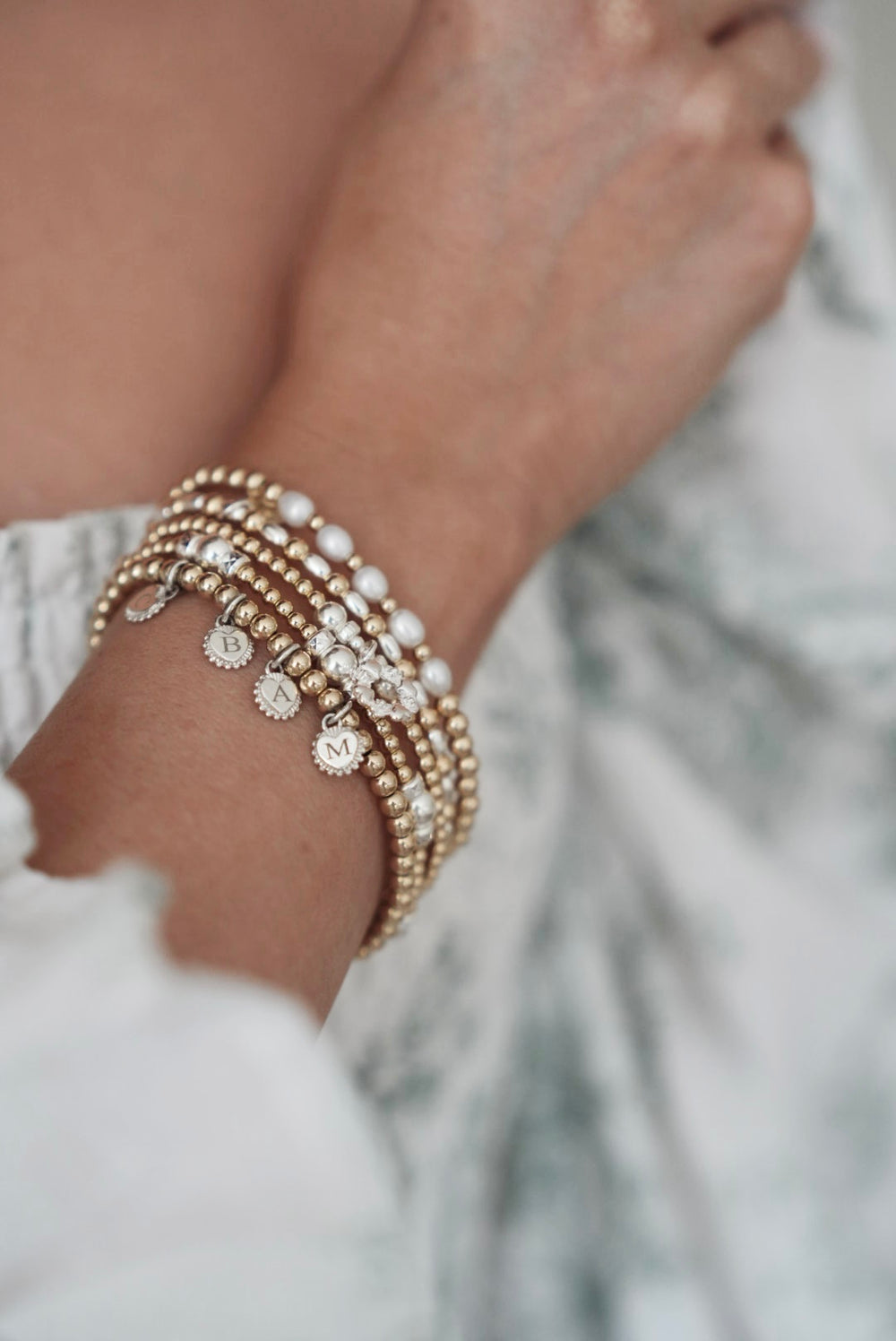 Close-up of a wrist wearing multiple gold, silver and pearl beaded stacking bracelets with blurred background
