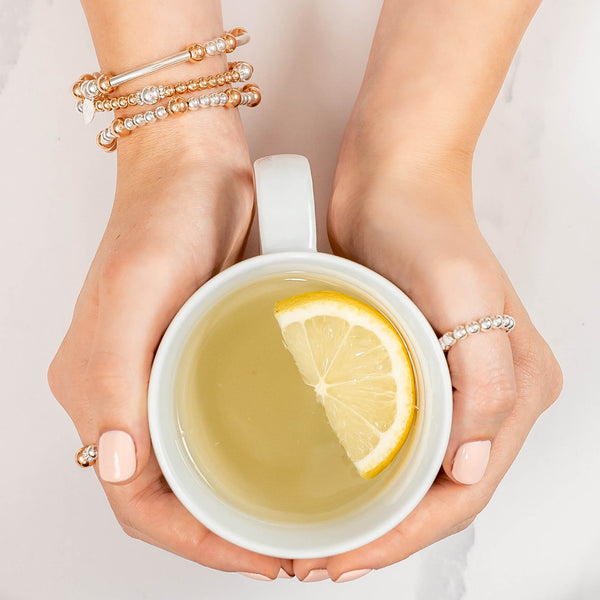 Hands holding a cup of tea with a lemon slice, wearing silver and rose gold beaded stacking bracelets and rings on a light background