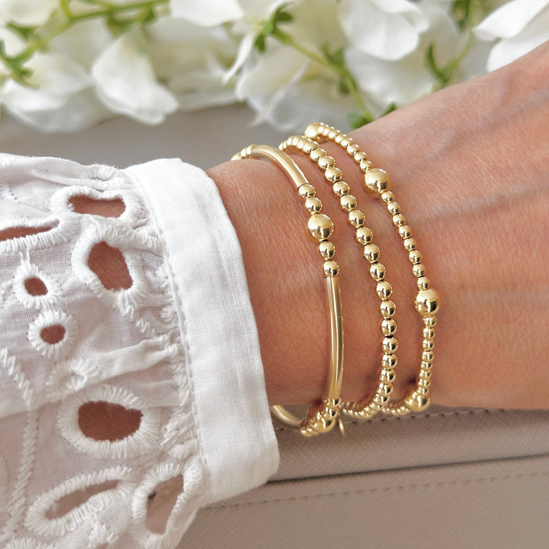 An aesthetic shot of gold beaded stacking bracelets on a wrist with some blurry flowers in the backround
