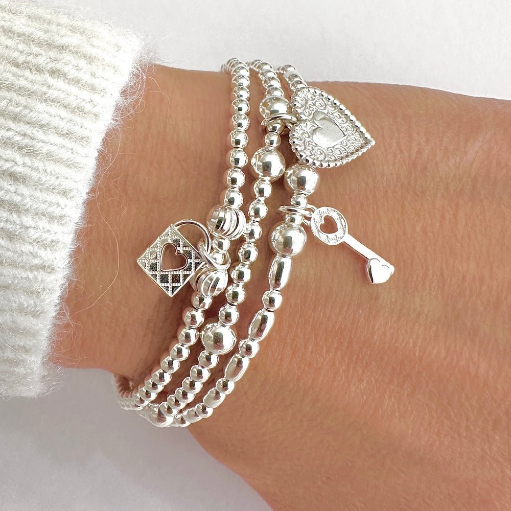 Three silver bracelets with heart, padlock and key charms on a wrist against a white background