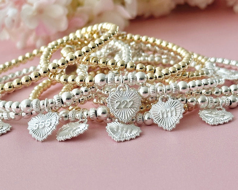 Stack of gold and silver beaded bracelets with angel number heart charms on a pink background