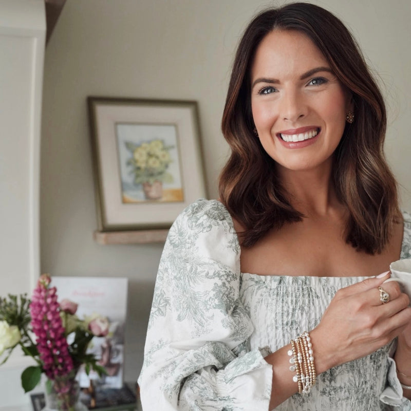 A close-up of Anna Corry (aka @blossomingbirds on Instagram) holding a mug in a cosy indoor setting with flowers and a painting in the background, wearing a stack of gold and silver mixed-metal beaded Bella Jane stacking bracelets.