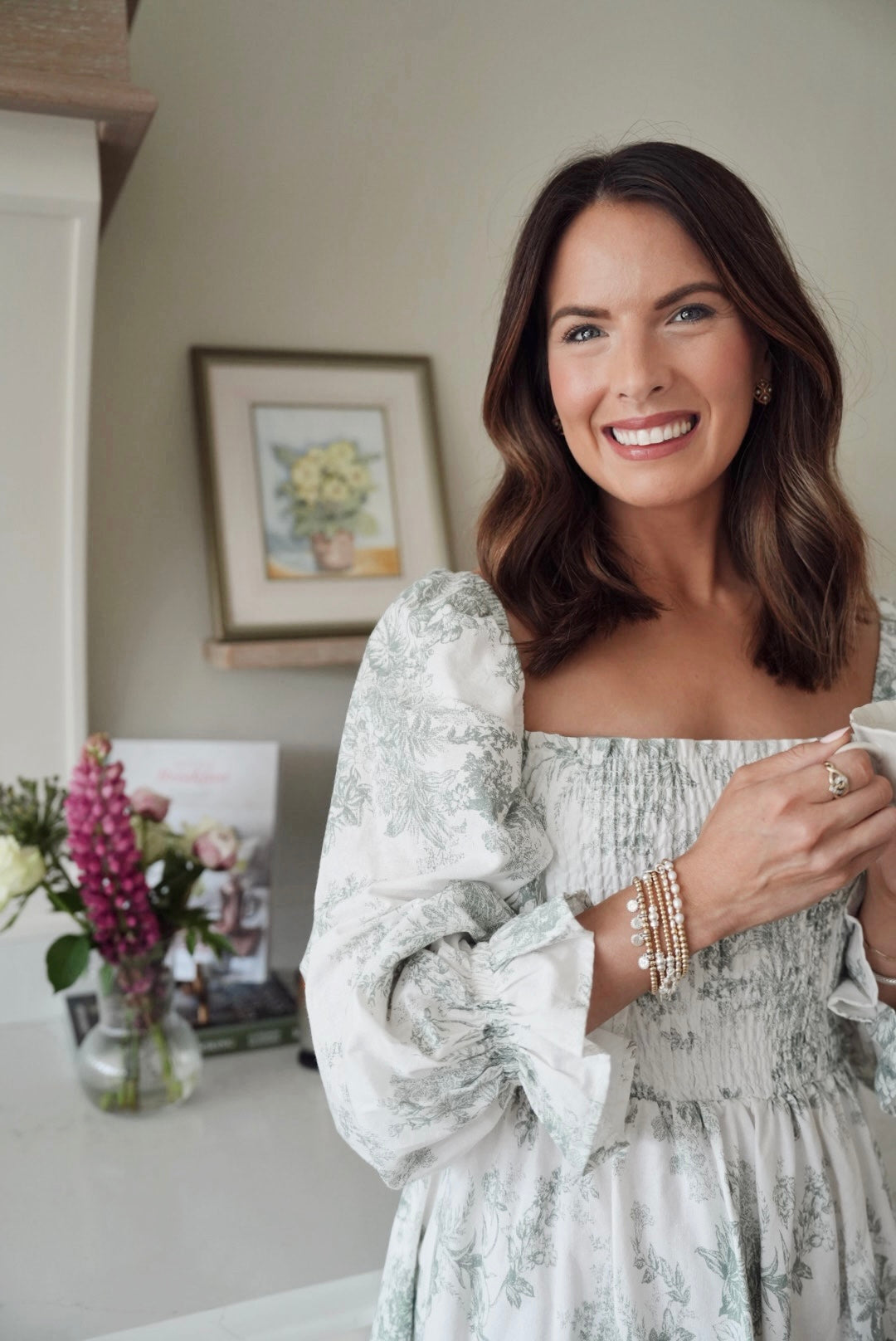 A close-up of Anna Corry (aka @blossomingbirds on Instagram) holding a mug in a cosy indoor setting with flowers and a painting in the background, wearing a stack of gold and silver mixed-metal beaded Bella Jane stacking bracelets.