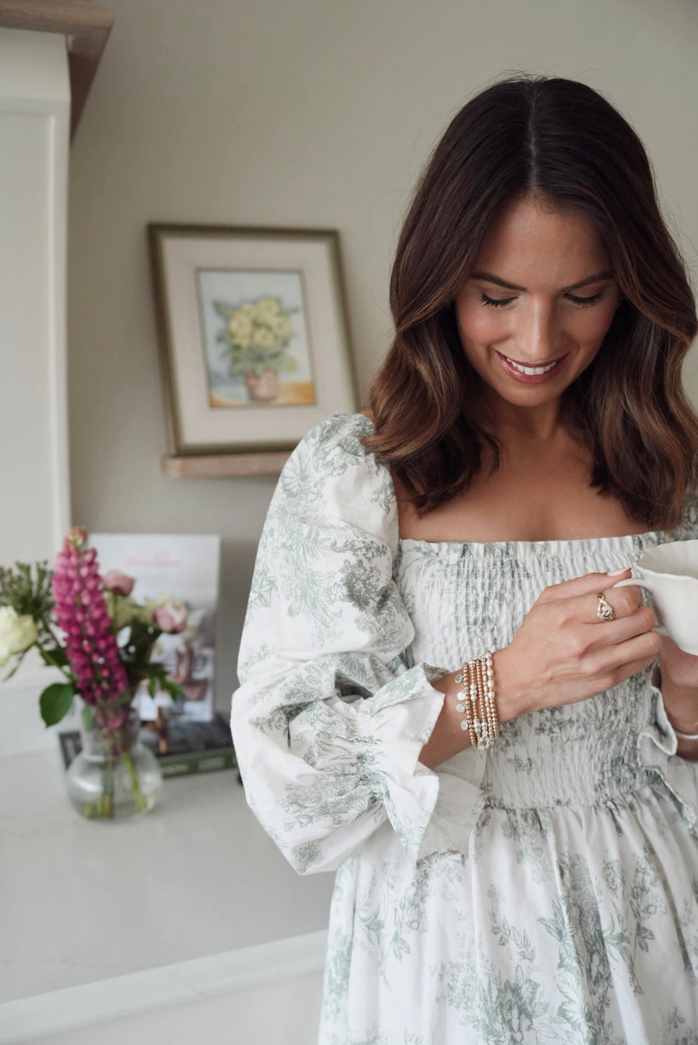 A close-up of Anna Corry (aka @blossomingbirds on Instagram) holding a mug in a cosy indoor setting with flowers and a painting in the background, wearing a stack of gold and silver mixed-metal beaded Bella Jane stacking bracelets.