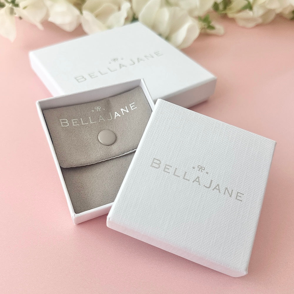 Bella Jane branded small grey pouch with white box on a pink background with white flowers in the background.
