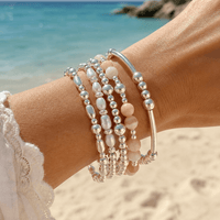 A close-up of a person's wrist wearing a stack of beaded bracelets on a sandy beach with the ocean in the background. The bracelets feature sterling silver beads, white pearl-like beads, and pale yellow coloured beads.