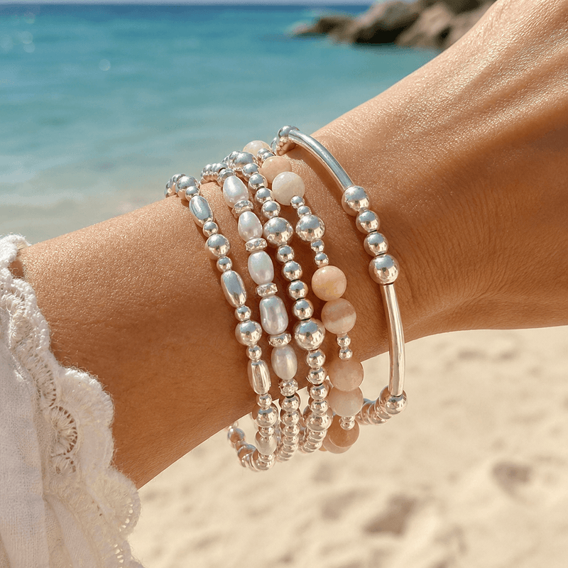 A close-up of a person's wrist wearing a stack of beaded bracelets on a sandy beach with the ocean in the background. The bracelets feature sterling silver beads, white pearl-like beads, and pale yellow coloured beads.
