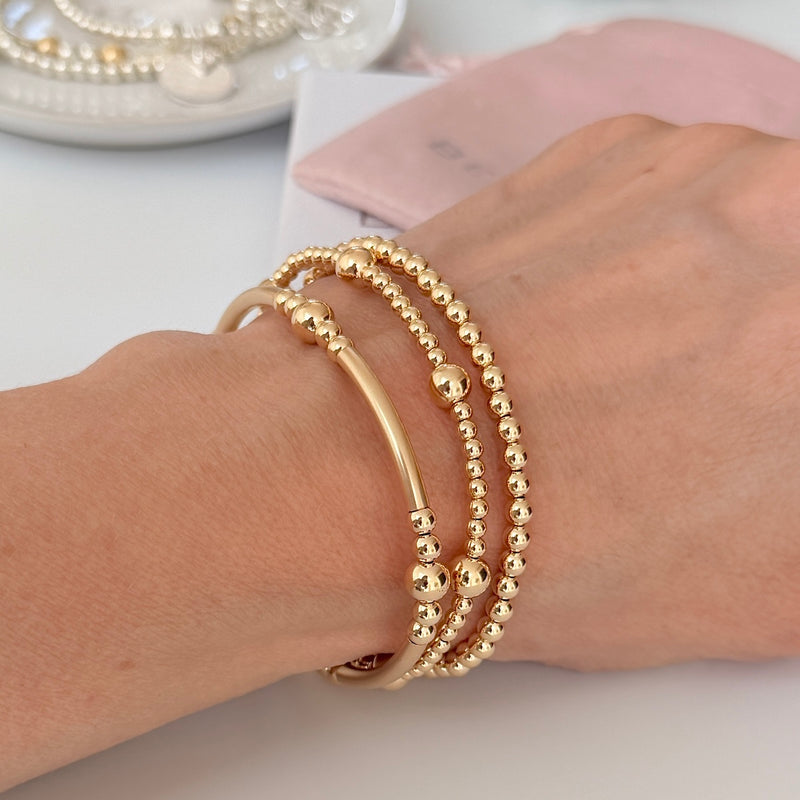 A close-up of a person's wrist wearing three layered gold bracelets: one noodle and bead shaped bracelet, one small round beaded bracelet, and one beaded bracelet with smaller beads and larger accent beads. A soft pink jewellery pouch and plate of bracelets are blurred in the background.