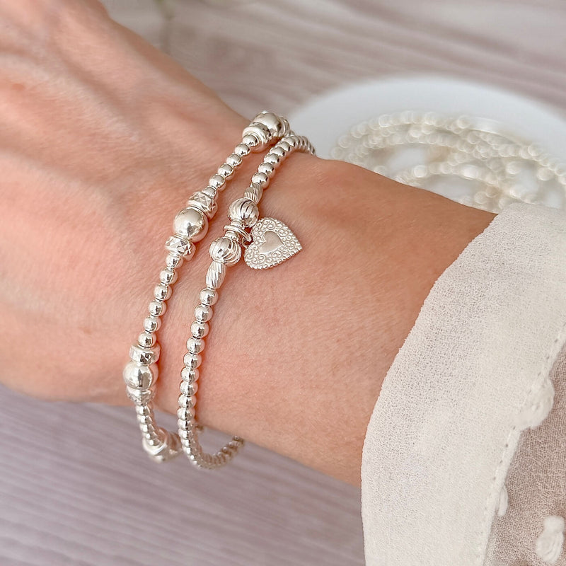 2 Silver beaded bracelets, one with a small heart charm on a wrist against a neutral background