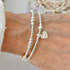 Silver beaded bracelet duo with a delicate heart charm on a wrist against a neutral background
