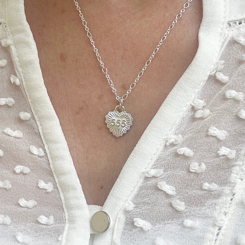 A close up of a model wearing a cream blouse with silver fine chain, featuring a a heart pendant with the angel number 555.
