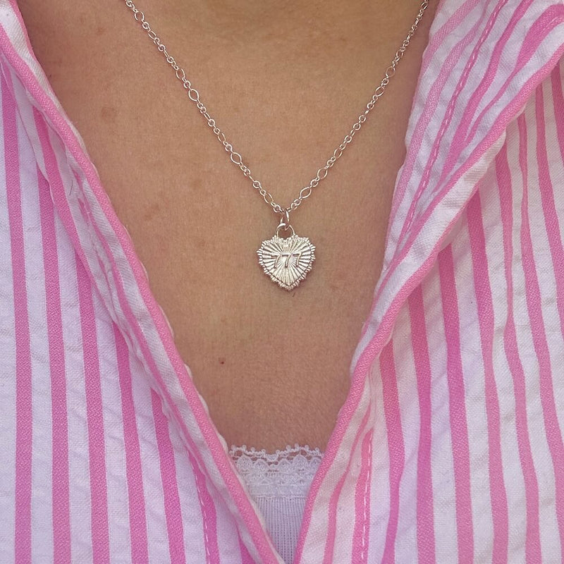 A close up of a a model wearing a silver necklace with a heart pendant featuring angel number 777. The model is wearing a pink and white striped shirt.