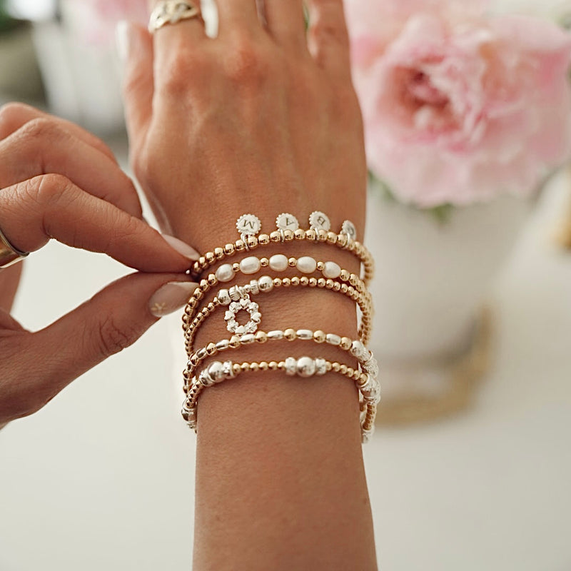 Close-up of Anna Corry's hand wearing multiple gold, silver and pearl beaded bracelets with a blurred floral background.