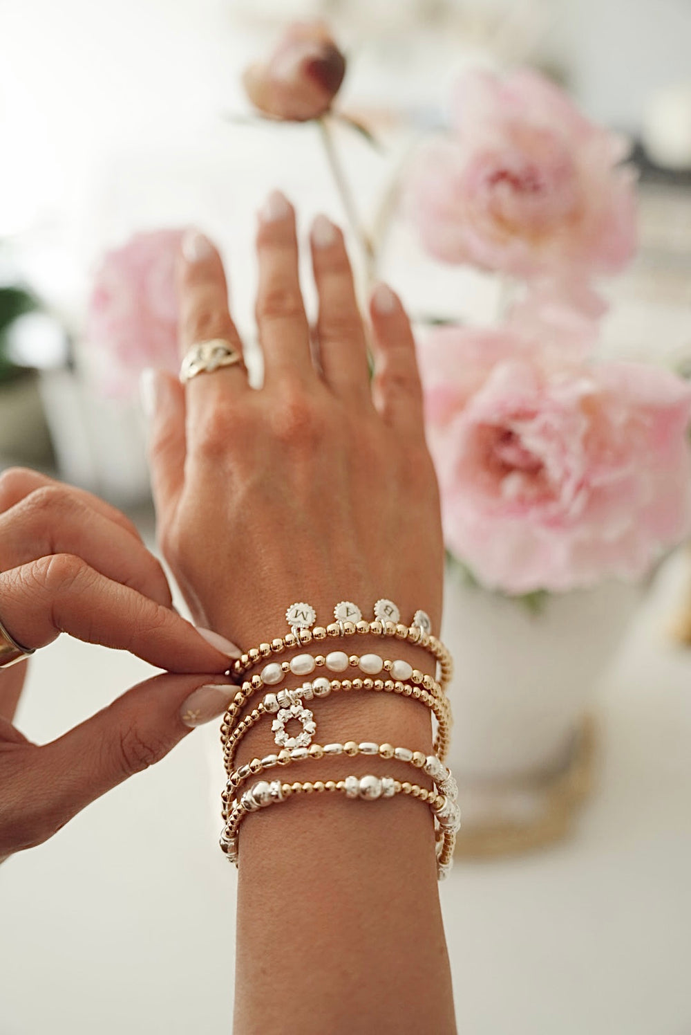 Close-up of Anna Corry's hand wearing multiple gold, silver and pearl beaded bracelets with a blurred floral background.