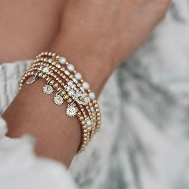 Close-up of a wrist wearing multiple gold, silver and pearl beaded stacking bracelets with blurred background
