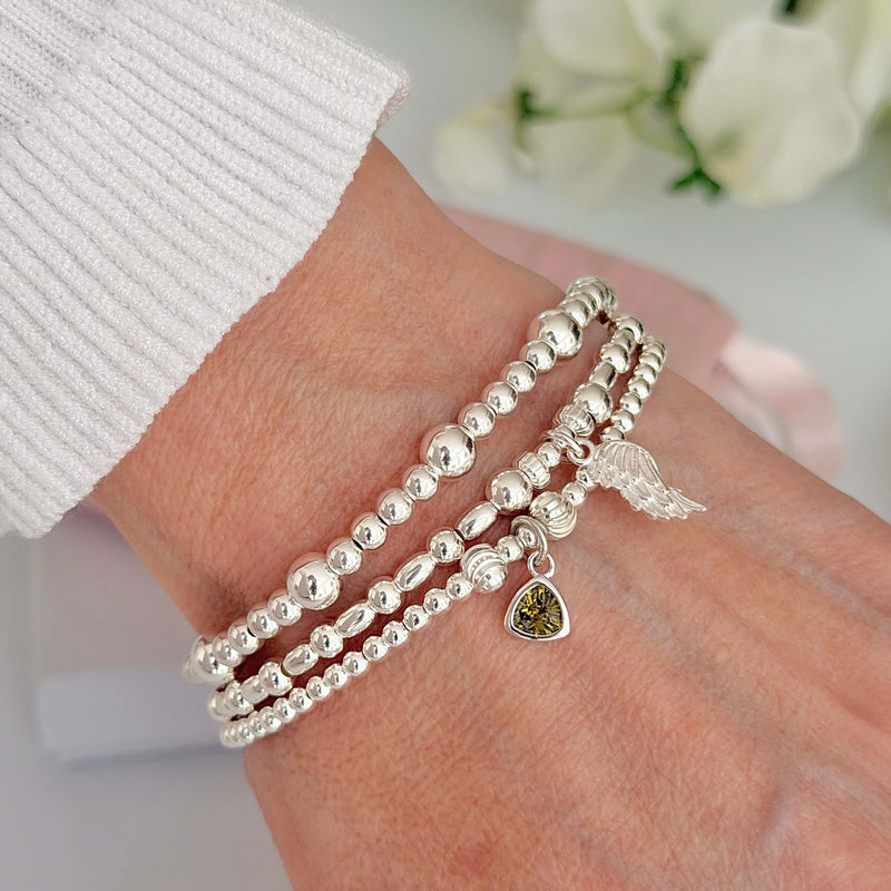 Silver beaded bracelets on a wrist, one with the August birthstone charm, against a blurred background of white flowers