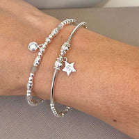 A close up of 2 silver beaded stacking bracelets, one featuring a small star charm with a tiny cubic zirconia stone in centre of the charm and another featuring pale grey coloured beads with a sparkly round charm, modelled on a wrist against a neutral background