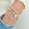 A close up of a lady's wrist with 5 silver stacking bracelets, one bracelet featuring pearls and a silver heart charm, and other featuring the April (diamond) birthstone charm. The lady has her hand in a jeans pocket.