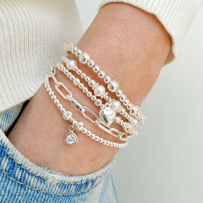 A close up of a lady's wrist with 5 silver stacking bracelets, one bracelet featuring pearls and a silver heart charm, and other featuring the April (diamond) birthstone charm. The lady has her hand in a jeans pocket.