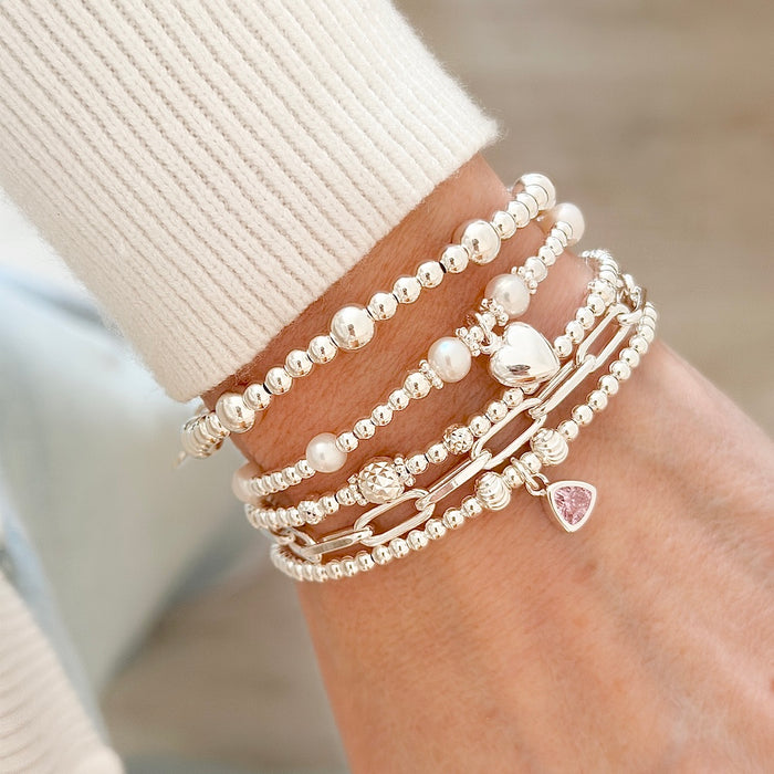 Close-up of a wrist wearing multiple silver beaded stacking bracelets one with a silver heart charm and another with pink October birthstone against a light background.