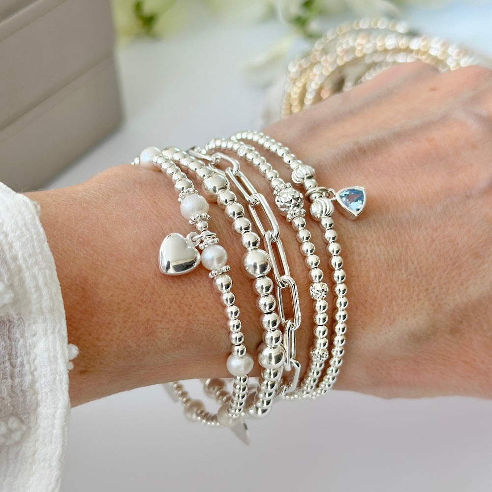 Close-up of a wrist wearing multiple silver beaded stacking bracelets with heart and pearl designs on a light background.