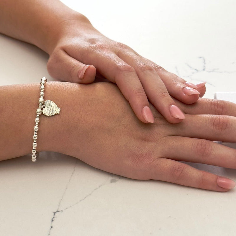 Close-up of a hand wearing a bracelet with a cherish charm which is a angel wings shaped into a heart on a marble surface