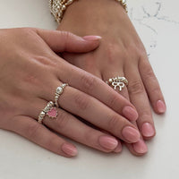 Two hands wearing silver rings with a pink  heart and small bow charms on a light background