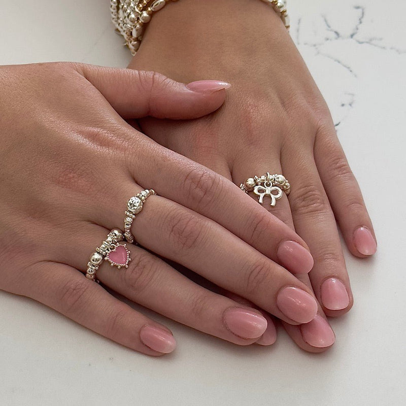 Two hands wearing silver rings with a pink  heart and small bow charms on a light background