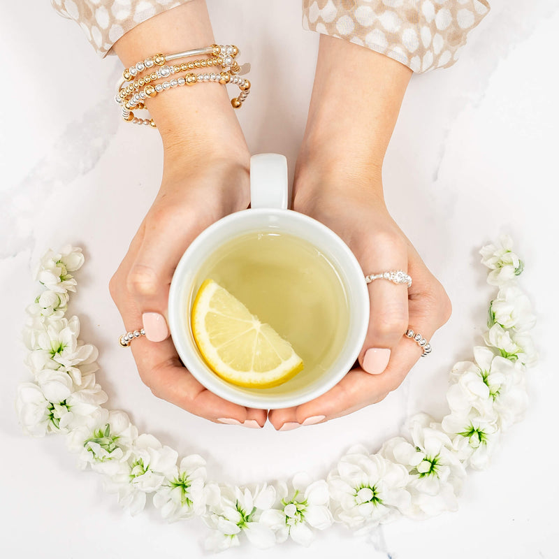 Hands holding a cup of tea with a lemon slice, showing a wrist wearing 3 silver and gold mixed metal stacking bracelets, surrounded by white flowers