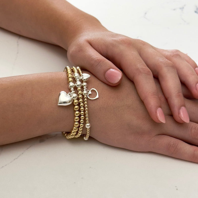 Close-up of a hand wearing multiple gold and silver mixed metal heart charm stacking bracelets on a light background