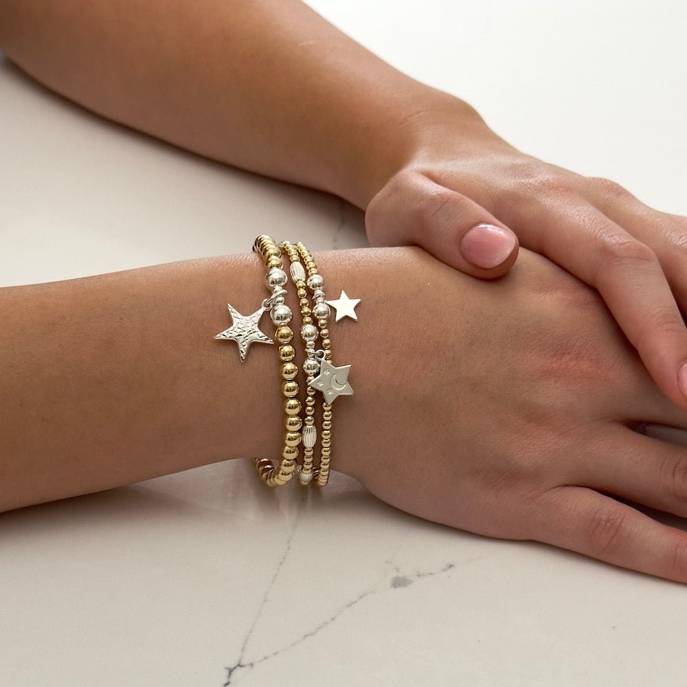 Close-up of a hand wearing multiple gold and silver mixed metal star charm stacking bracelets on a light background