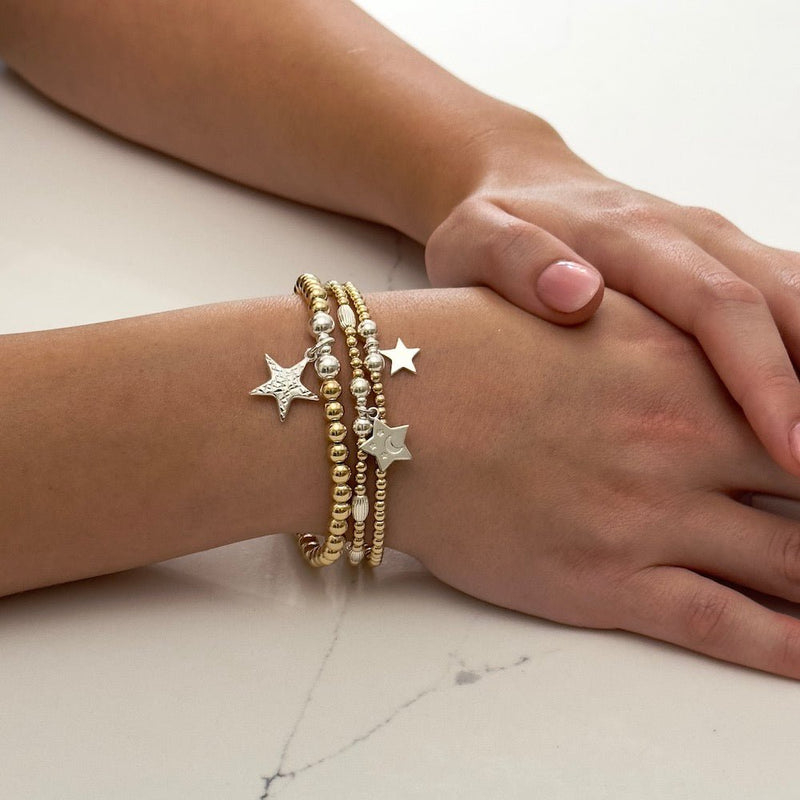 Close-up of a hand wearing multiple gold and silver mixed metal star charm stacking bracelets on a light background