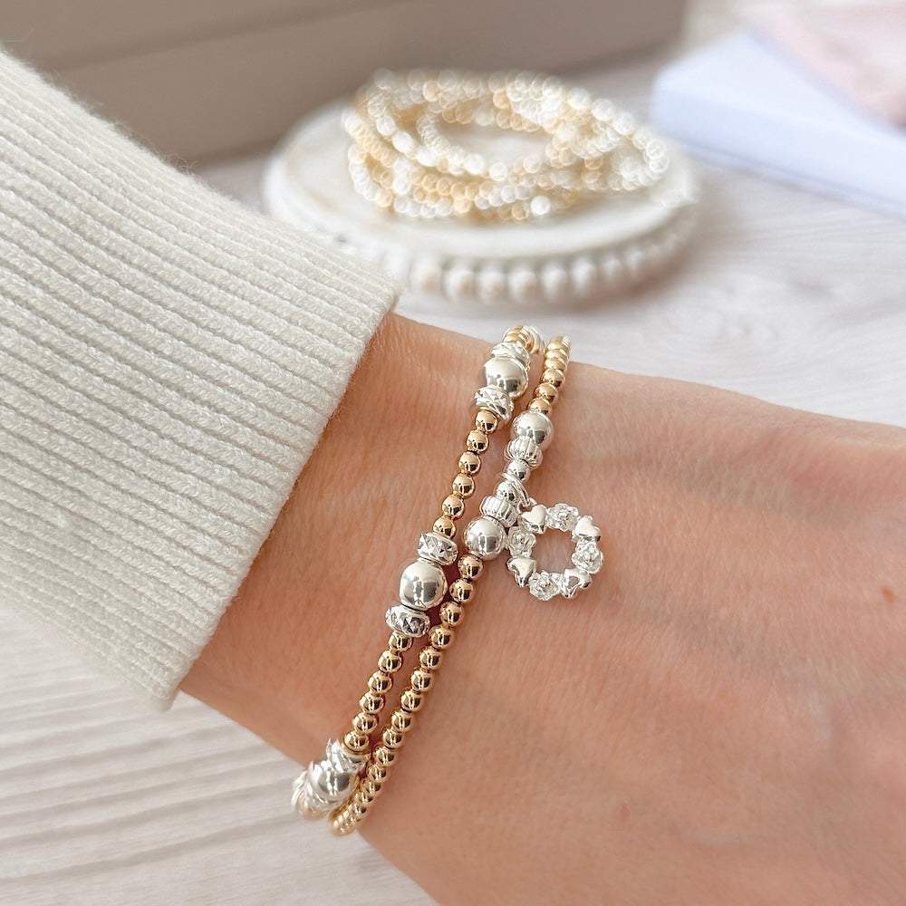 Close up of two gold and silver mixed metal beaded stacking bracelets on a wrist, one featuring a sterling silver wreath shaped charm, with a pink cream sleeve and bracelet stack in the background.