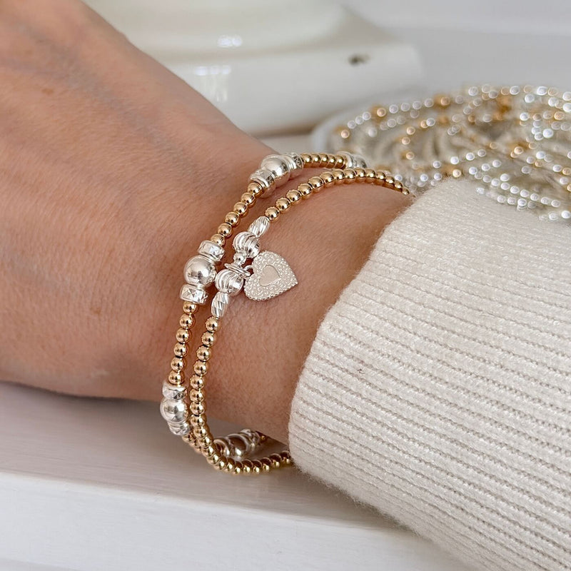 Close-up of a wrist wearing gold and silver beaded bracelets, one featuring a silver heart charm, on a light background