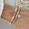 Close-up of a wrist wearing two beaded gold and silver mixed metal bracelets, with heart charm on a neutral background