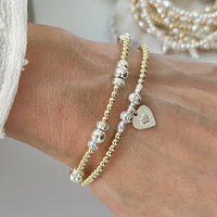 Close-up of a wrist wearing two beaded gold and silver mixed metal bracelets, with heart charm on a neutral background