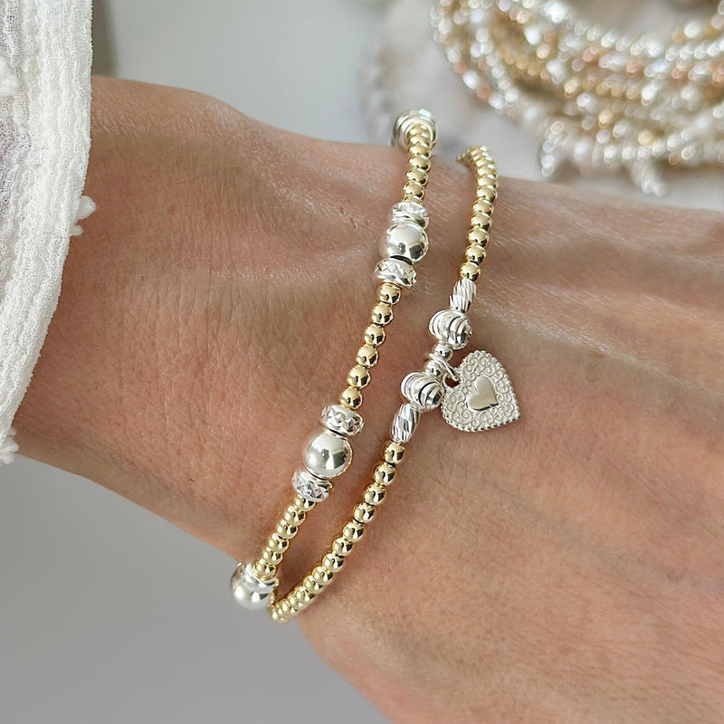 Close-up of a wrist wearing two beaded gold and silver mixed metal bracelets, with heart charm on a neutral background