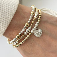 A close up photo of a woman's wrist, featuring 3 sterling silver and gold-filled beaded stacking bracelets, one with a sterling silver heart-shaped charm featuring angel number 222 on the front of the charm.