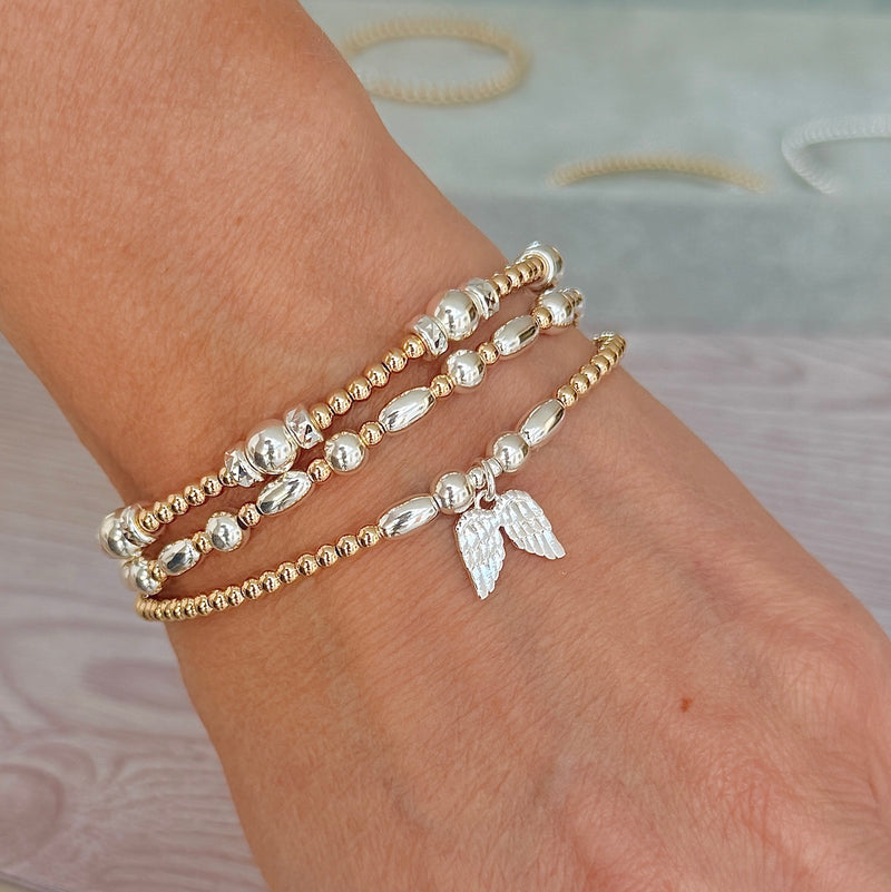 A close-up of a person's wrist wearing three silver and gold mixed-metal stacked beaded bracelets, one with a silver angel wings charm.