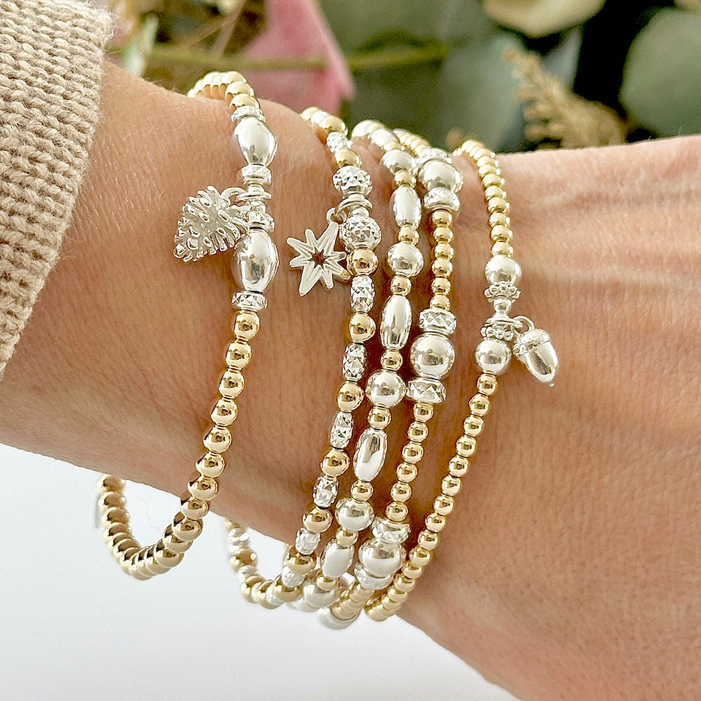 Close-up of a wrist wearing multiple silver and gold mixed metal stacking bracelets, with a silver pine cone, acorn and star charms, on a neutral background