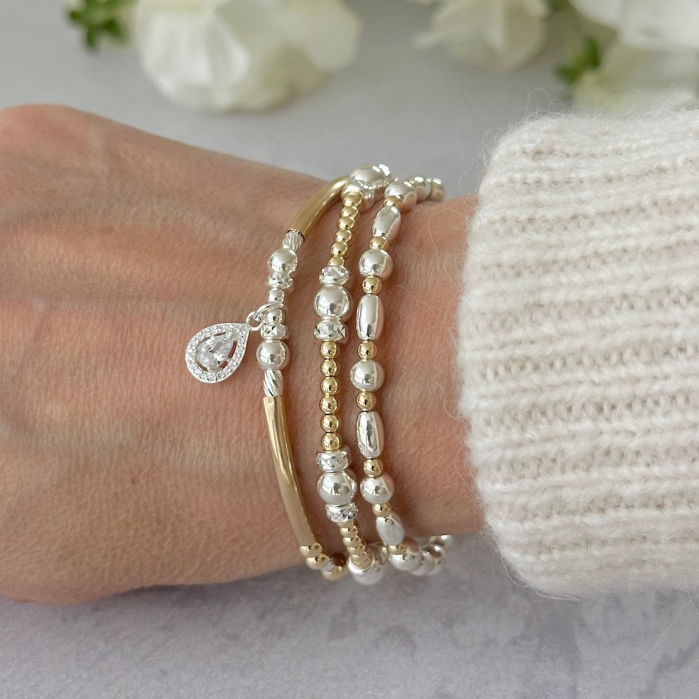 Close-up of a wrist wearing multiple beaded gold and silver mixed metal stacking bracelets, one with a silver and cubic zirconia tear drop charm, against a neutral background