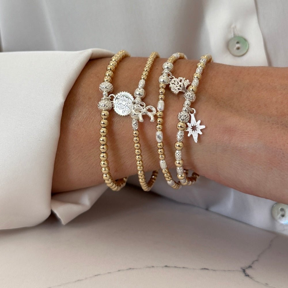 Close-up of a wrist wearing multiple gold and silver mixed metal beaded bracelets with various charms on a neutral background.