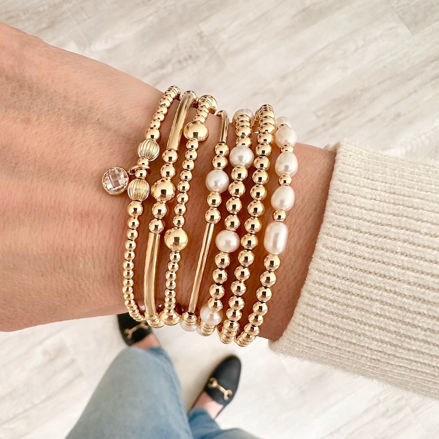 A model standing up with a close up of her wrist that is wearing a mix of handmade, beaded, gold-filled stacking bracelet. One features a cubic zirconia birthstone charm, three bracelets feature freshwater pearls and the rest are plain gold