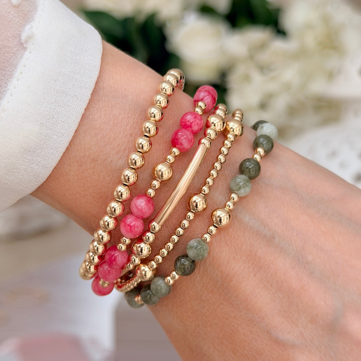 A close-up of a person's wrist wearing multiple beaded bracelets, including gold-filled beads, pink stone beads, and olive green stone beads, with a white sleeve visible at the left edge.