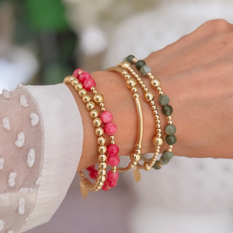 A close-up of a person's wrist wearing multiple beaded bracelets, including pink and green stone beads with gold-filled  beads, against a softly blurred light background.