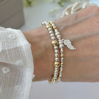 Silver and gold mixed metal bracelet duo with angel wing charm on a wrist, with a blurred image of stacking bracelets in the background.