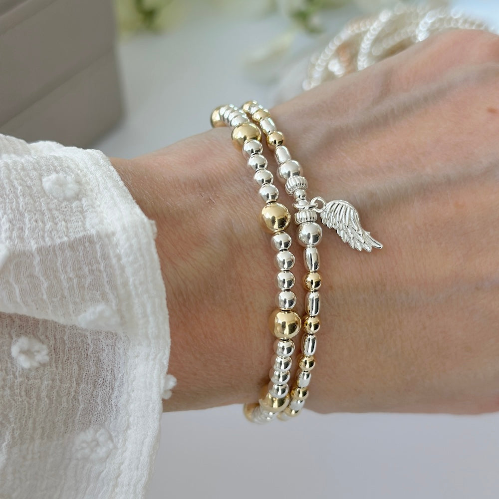 Silver and gold mixed metal bracelet duo with angel wing charm on a wrist, with a blurred image of stacking bracelets in the background.
