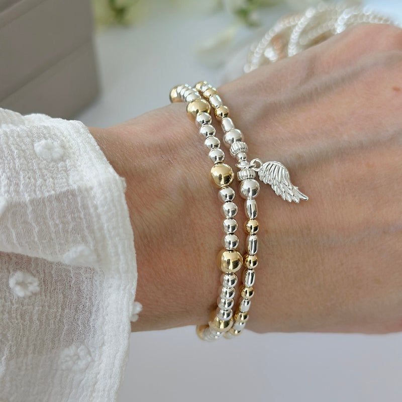 Silver and gold mixed metal bracelet duo with angel wing charm on a wrist, with a blurred image of stacking bracelets in the background.