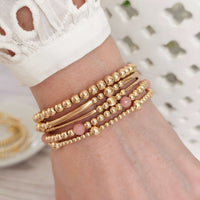 Gold-filled beaded stacking bracelets with one gold and pink opal stacking bracelet, on a wrist with a blurred neutral background